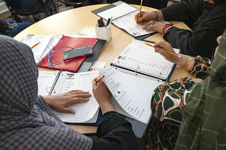 A round table full of literacy workbooks with three refugee women sitting around it.